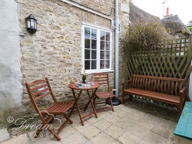 An outdoor patio with a small wooden table, two wooden chairs, and a wooden bench against a stone wall at Bramble Cottage in Burton Bradstock