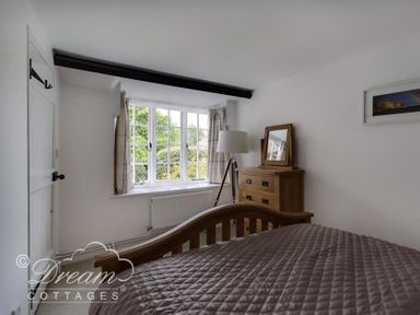 A bedroom with a wooden bed, wooden dresser with mirror, floor lamp and window with curtains at Bramble Cottage in Burton Bradstock