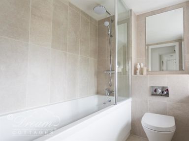 A bathroom with a bathtub shower combination a wall-mounted toilet and a large mirror at Bramble Cottage in Burton Bradstock