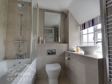 A bathroom with a bathtub shower combination a toilet a sink on a tiled counter and a window with a striped valance at Bramble Cottage in Burton Bradstock