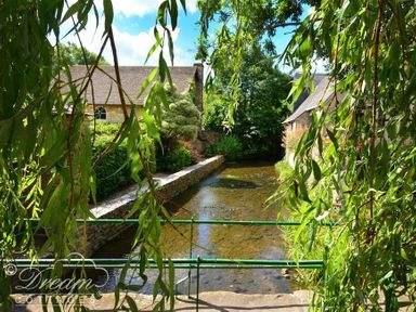A small river flowing between stone houses with greenery and overhanging tree branches at Bramble Cottage in Burton Bradstock