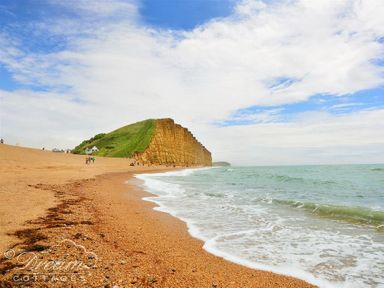 A pebble beach with waves and a green cliff in the background at Bramble Cottage in Burton Bradstock
