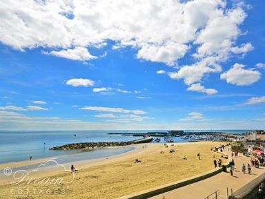 A sandy beach with people walking and sitting near a harbor with boats under a blue sky with clouds at Bramble Cottage in Burton Bradstock
