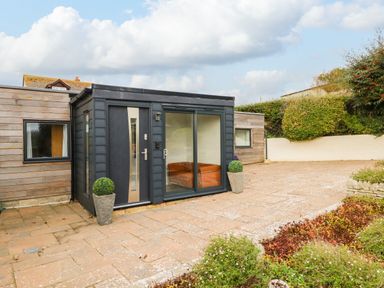 A patio area with potted plants and a wooden building with a black door and large glass window at Burton Bay in Burton Bradstock