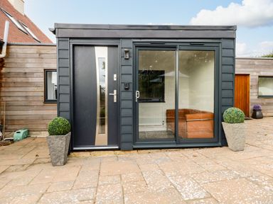 A small modern building with a black door and glass sliding door between two potted plants on a paved patio at Burton Bay in Burton Bradstock