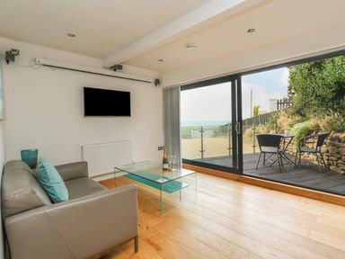 A living room with a gray sofa and glass coffee table facing a mounted TV and sliding glass doors at Burton Bay in Burton Bradstock