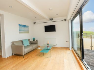 A living room with a sofa a glass coffee table a wall-mounted tv and large sliding glass doors at Burton Bay in Burton Bradstock