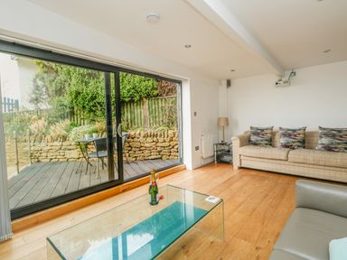 A living room with a glass coffee table a beige sofa with three patterned cushions a floor lamp a projector and large sliding glass doors leading to an outdoor seating area at Burton Bay in Burton Bradstock