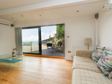 A living room with a beige sofa and glass coffee table next to sliding glass doors leading to an outdoor patio at Burton Bay in Burton Bradstock