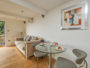 A small dining area with a glass round table and two gray chairs beside a beige sofa with three cushions and a wall painting at Burton Bay in Burton Bradstock