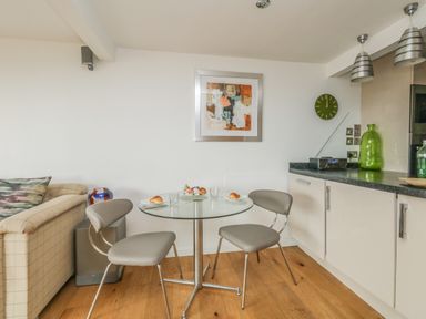 A dining area with a glass table and two chairs next to a kitchen counter with cabinets and a clock on the wall at Burton Bay in Burton Bradstock