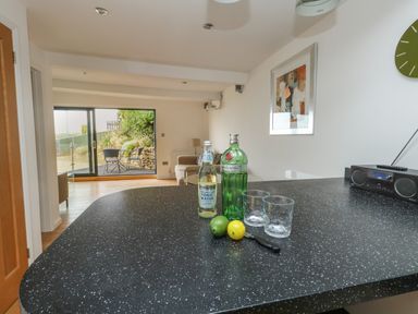 A kitchen countertop with bottles glass cups lime lemon and a knife a living area with sofa and glass sliding door at Burton Bay in Burton Bradstock