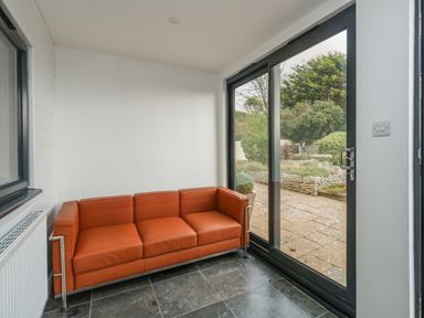 A room with a brown sofa next to a window and glass sliding doors showing a garden outside at Burton Bay in Burton Bradstock