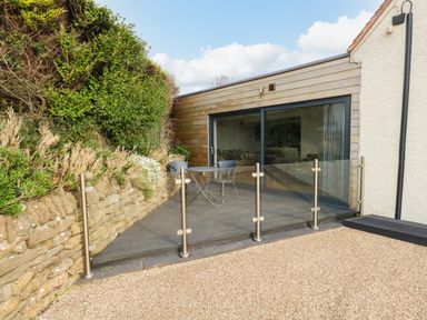 A patio area with a small round table and two chairs behind glass fencing next to a building with sliding glass doors and a stone wall with plants at Burton Bay in Burton Bradstock