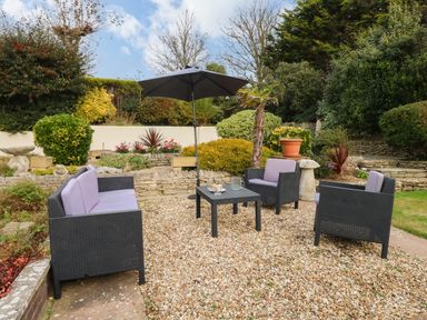 An outdoor seating area with black wicker furniture and purple cushions around a black table with an umbrella in a garden at Burton Bay in Burton Bradstock