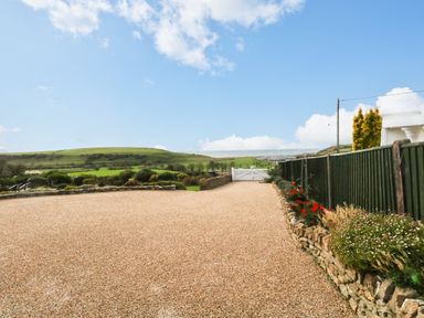 A gravel driveway with a green fence and stone flower beds leading to a white gate with hills and sky in the background at Burton Bay in Burton Bradstock