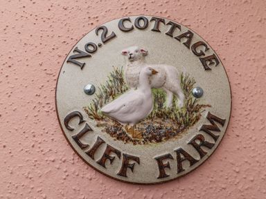 A circular sign on a pink textured wall with a lamb and a goose and text saying No 2 Cottage Cliff Farm