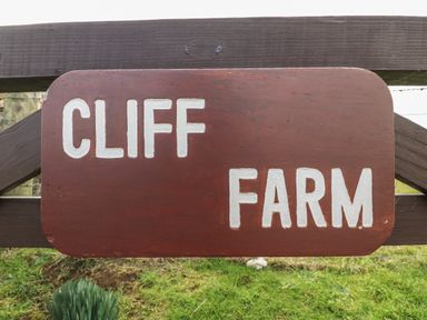 A wooden sign with white letters reading Cliff Farm mounted on a wooden fence with grass in the background at Cliff Farm No 2 Cottage in Burton Bradstock