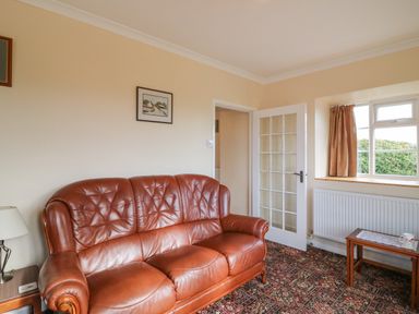 A living room with a brown leather sofa a wooden side table with a lamp a small wooden table near window and a door with glass panels at Cliff Farm No 2 Cottage Burton Bradstock