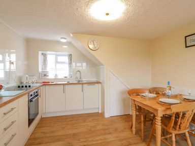 A kitchen with white cabinets a stove and oven a wooden dining table with four chairs and a clock on the wall at Cliff Farm No 2 Cottage Burton Bradstock