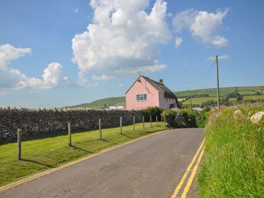 A rural road bordered by grass and a stone wall with a pink house and green hills in the background at Cliff Farm No 2 Cottage Burton Bradstock