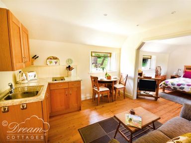 A small kitchen with wooden cabinets and a sink an adjoining dining area with a round table and two chairs then a bedroom with a bed a small TV and wooden furniture at Coombe Orchard in Swanage
