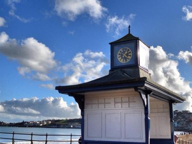 A small clock tower structure by the seaside with a railing and buildings in the background at Coombe Orchard in Swanage