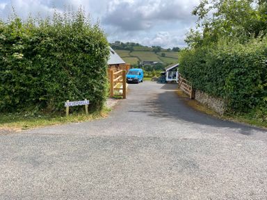A driveway entrance with hedges on both sides a blue van and a small building and fields in the background at Culverwells in Axminster