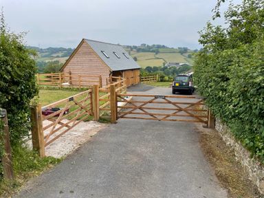 A driveway with wooden gates leading to a wooden garage and a car with rural fields in the background at Culverwells Axminster
