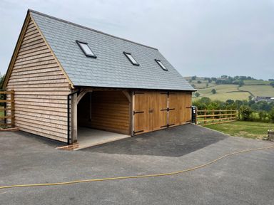 A wooden garage with a slate roof and three skylights next to a paved driveway in a rural landscape at Culverwells in Axminster