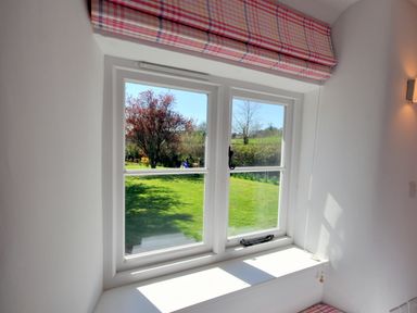 A window with a pink plaid roman blind showing a garden with grass trees and bushes at Culverwells in Axminster