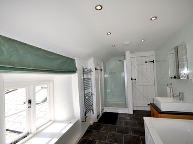 A bathroom with a window and green blind a heated towel rail a glass shower cubicle a sink with a mirror and a white wooden door at Culverwells in Axminster