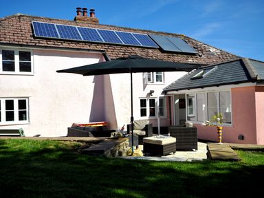 An outdoor patio area with chairs, a table, and an umbrella next to a pink house with solar panels on the roof at Culverwells in Axminster