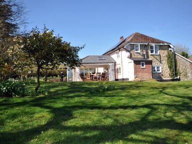 A garden with green grass trees and a patio with wooden chairs attached to a stone and brick house at Culverwells in Axminster