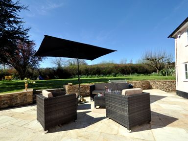 An outdoor patio with woven chairs and sofa under a large black umbrella with a garden and lawn in the background at Culverwells in Axminster