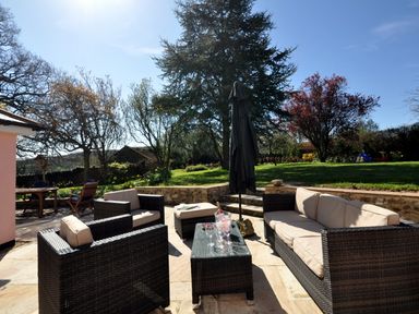 An outdoor patio with a wicker sofa set table and umbrella surrounded by grass trees and garden at Culverwells in Axminster