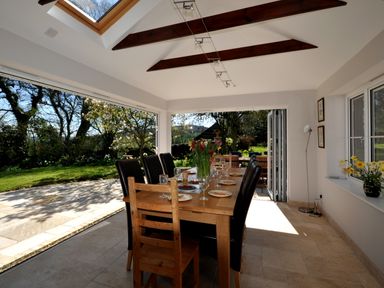 A dining room with a wooden table and leather chairs overlooking a garden through large open glass doors at Culverwells in Axminster