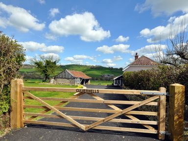 A wooden gate with the name Culverwells leading to a driveway with a house and barn with green fields and blue sky in the background at Culverwells in Axminster