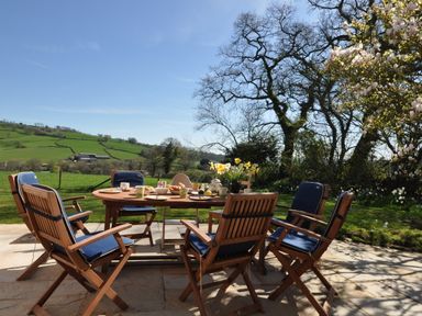 An outdoor patio with a wooden table and six chairs with blue cushions surrounded by trees and green fields at Culverwells in Axminster