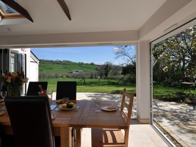 A dining area with a wooden table and chairs open to a garden and green hills at Culverwells in Axminster