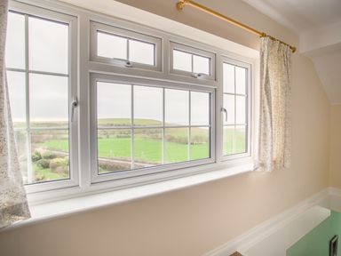 A window with white frames and floral curtains showing a green field view at The Doves in Burton Bradstock