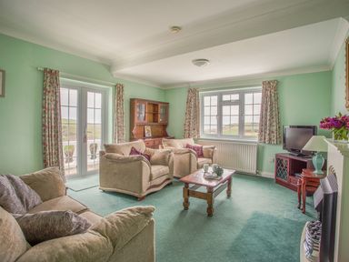 A living room with beige sofas a wooden coffee table a wooden cabinet a television and windows with floral curtains at The Doves in Burton Bradstock