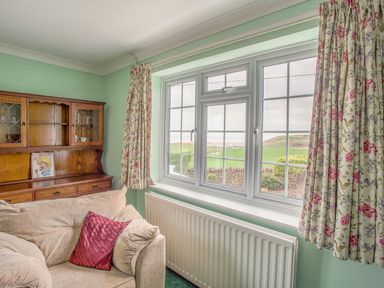 A sitting room with a beige sofa and floral curtains by a window with a countryside view at The Doves in Burton Bradstock