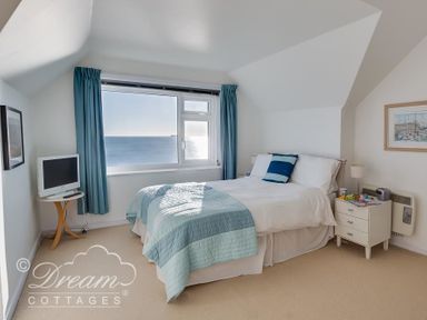 A bedroom with a bed beside a window showing a sea view with blue curtains and a small television on a wooden table at Dream View in Greenhill