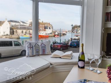 A window seat with a cushion and open book overlooking a harbor with boats and parked cars at Harbour Edge in Brewers Quay Harbour