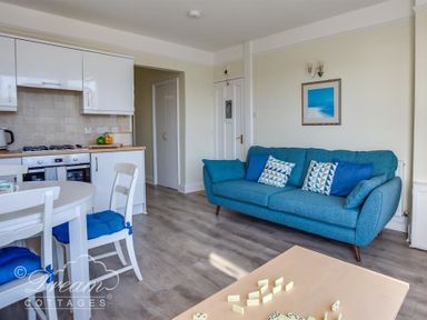 A living room and kitchen area with a blue sofa dining table with chairs and a coffee table with dominoes at Harbour Edge in Brewers Quay Harbour
