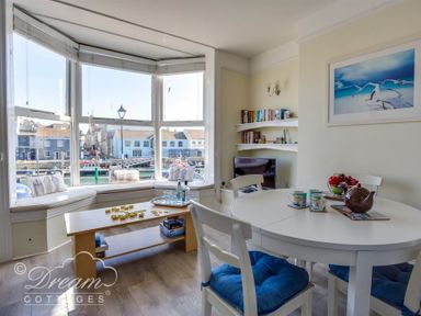 A dining area with a round white table and blue cushions on chairs beside a bay window with a view of harbor buildings at Harbour Edge in Brewers Quay Harbour