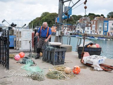 A man handling fishing nets on a dock with buoys ropes and boats in the water at Harbour Edge in Brewers Quay Harbour