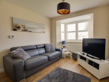 A living room with a grey leather couch and cushions a TV on a white stand and a bay window with blinds at The Ferns in Weymouth