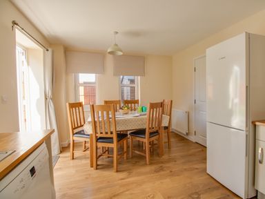 A dining area with a wooden table and six chairs next to windows and a white refrigerator at The Ferns in Weymouth